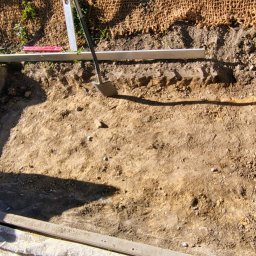 KSAL GREEN Pielęgnacja Ogrodów Kamil Kozioł - Wykopany, prostokątny dół w ziemi, przygotowany pod fundament ogrodzenia panelowego. Widoczna łopata, poziomica i taczka z kamieniami. Siatka ogrodzeniowa w tle.