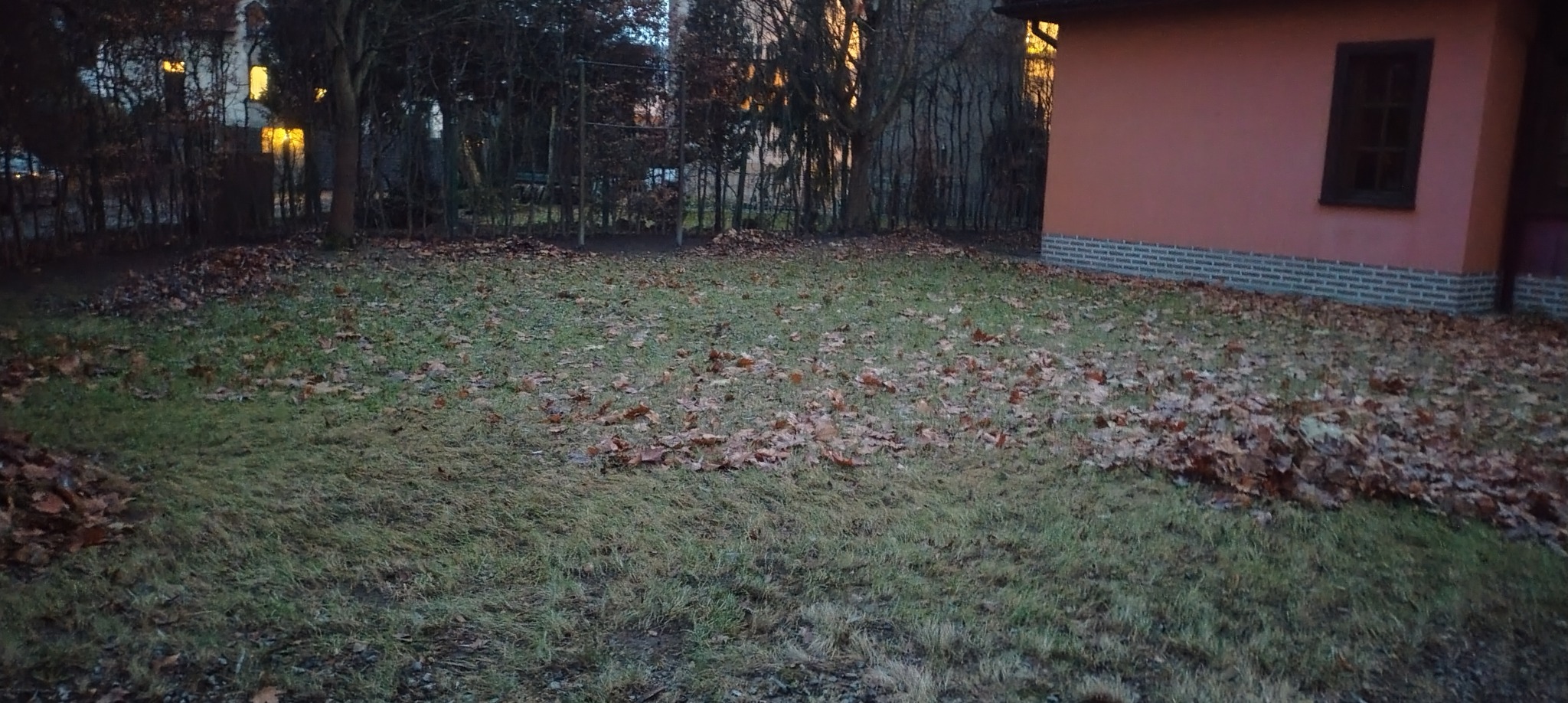 Jesienny trawnik z opadłymi liśćmi, częściowo zagrabionymi w pryzmę przy budynku. W tle drzewa i fragment placu zabaw.