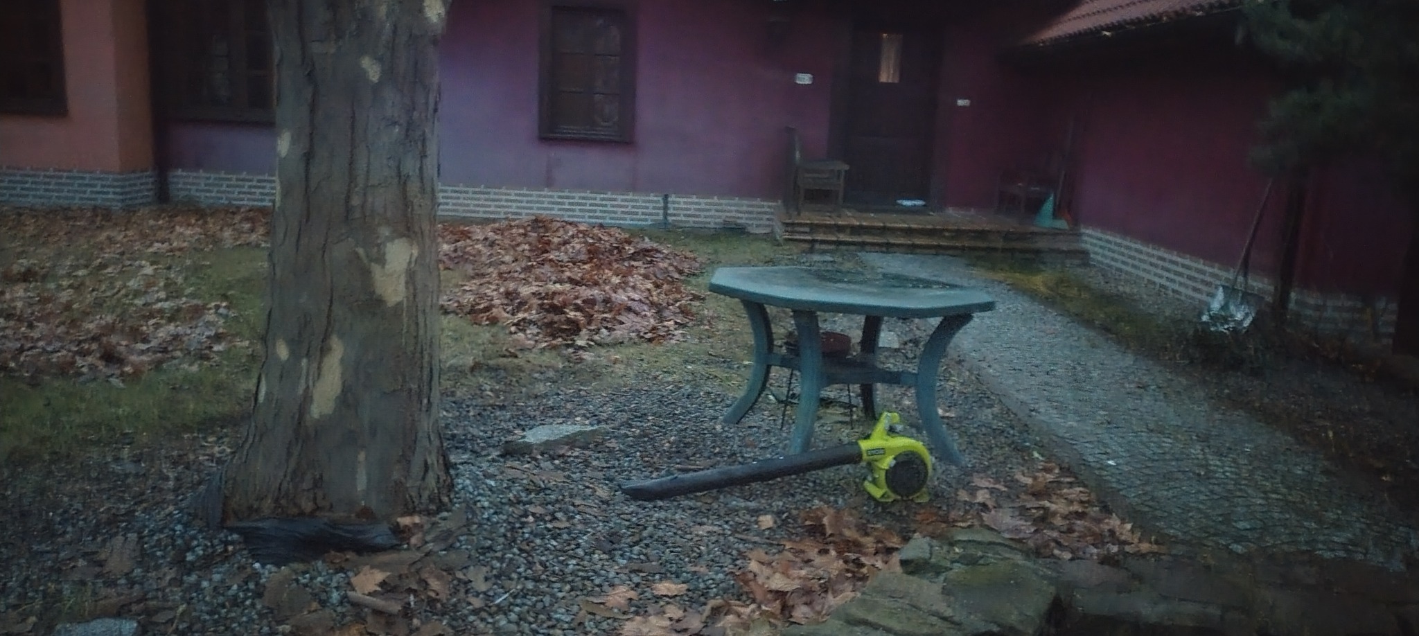 Sterta brązowych liści obok pnia drzewa, dmuchawa do liści leży na ziemi. W tle dom z ciemnymi oknami i drzwiami, oraz stół ogrodowy.