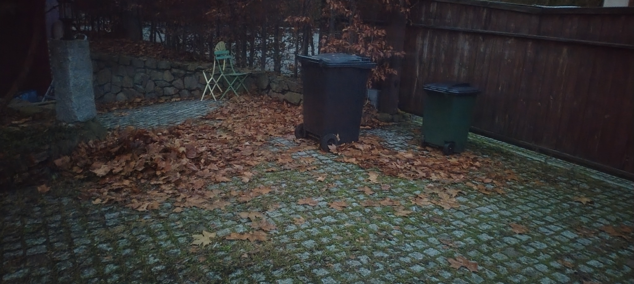 Jesienne liście na kostce brukowej, dwa pojemniki na śmieci, drewniany płot w tle. Sezonowe porządki w ogrodzie.
