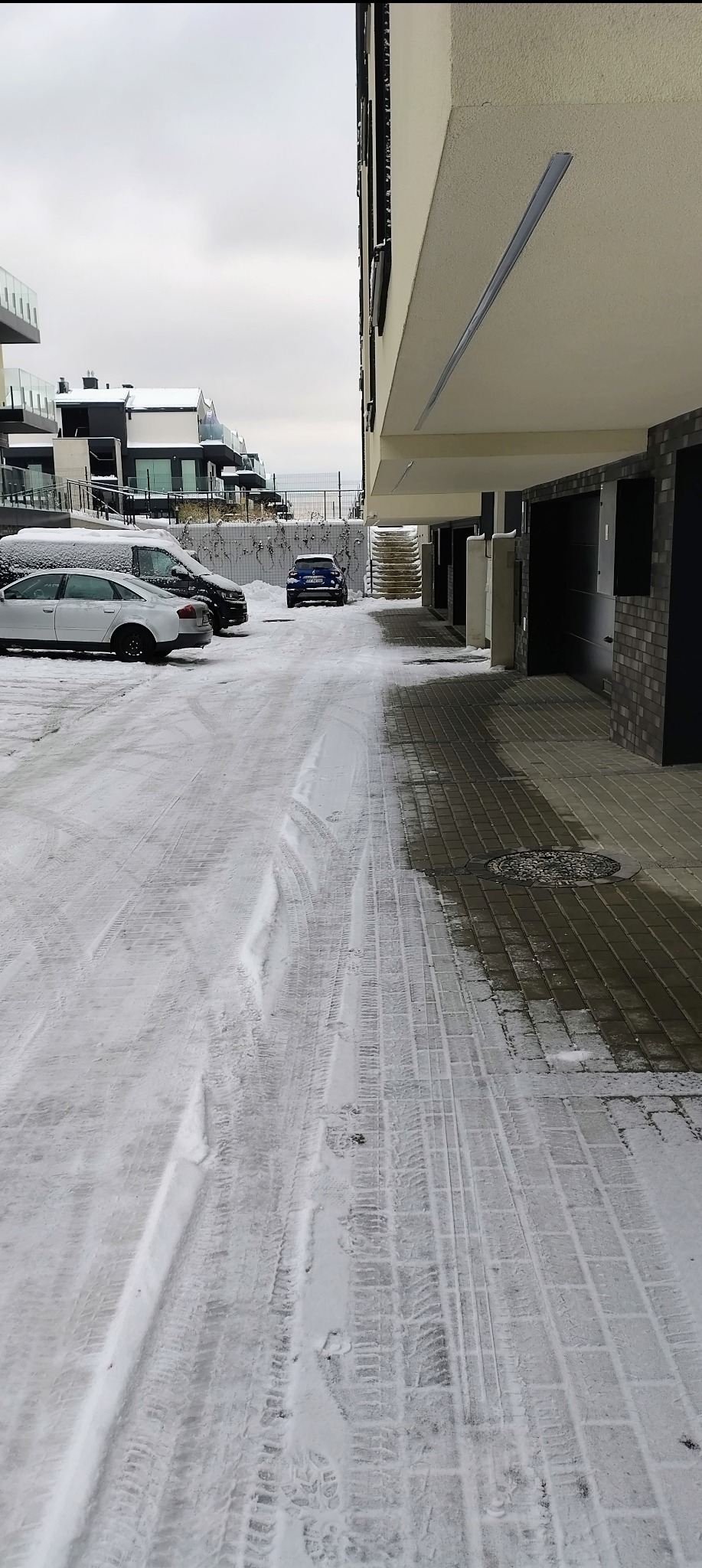 Zimowy widok na ośnieżony parking osiedlowy z widocznymi śladami opon, budynkami w tle i fragmentem podcieni. Pochmurne niebo.