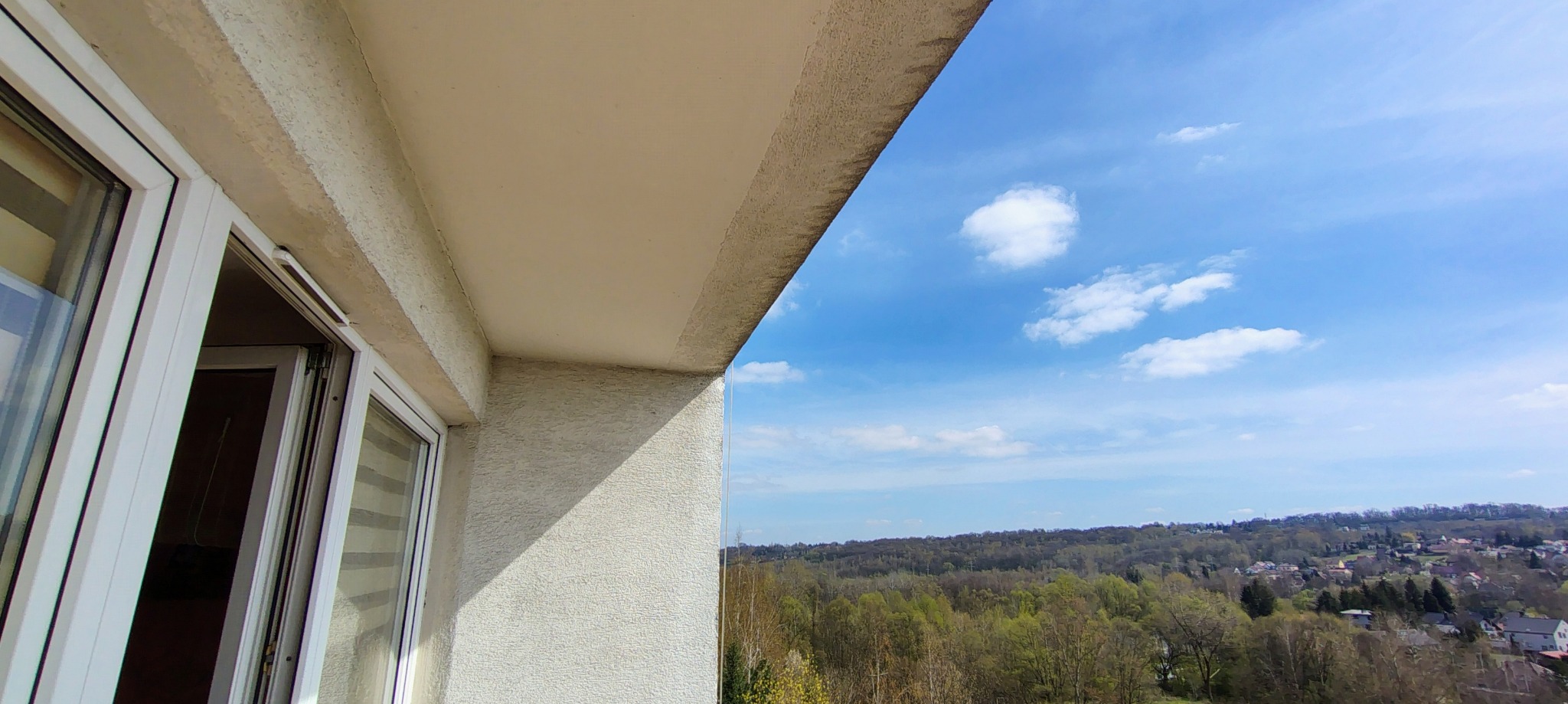 Widok z balkonu na zalesione wzgórza i błękitne niebo z chmurami. Częściowo widoczne białe ramy okienne i fragment elewacji budynku o jasnej, chropowatej fakturze.