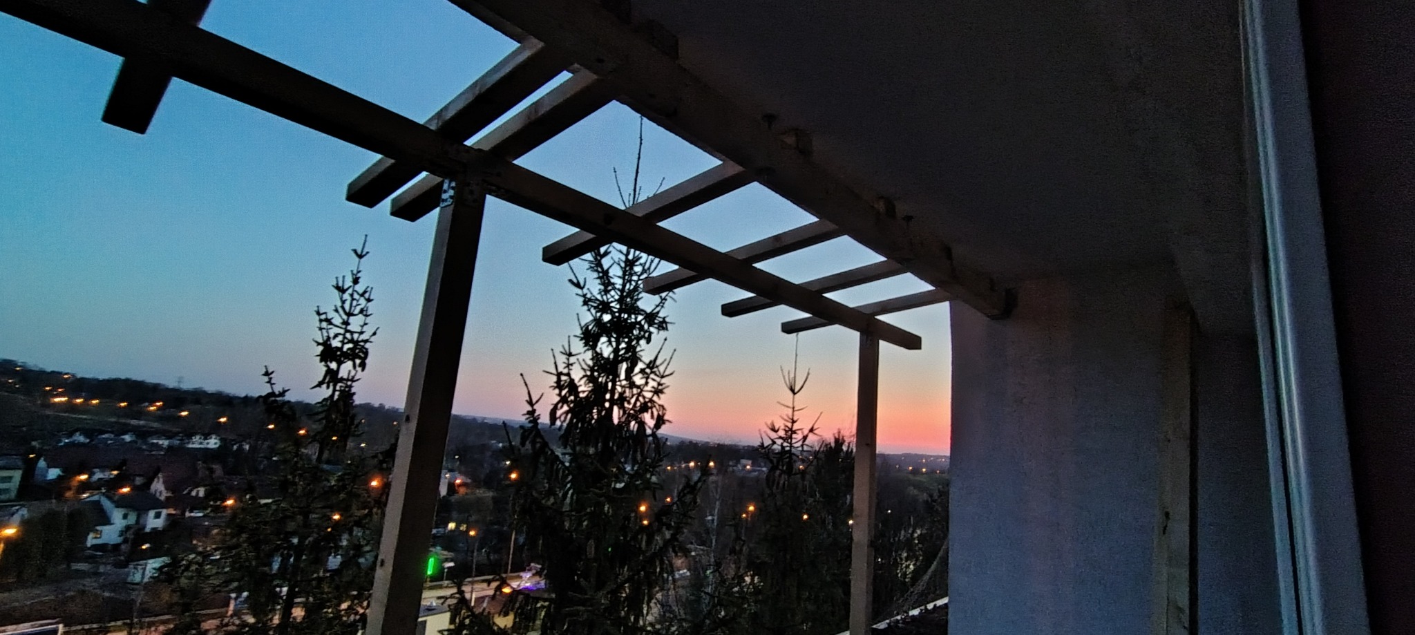 Drewniana konstrukcja pergoli na balkonie o zmierzchu, widok na miasto z delikatną poświatą zachodzącego słońca. Elementy konstrukcyjne z widocznymi mocowaniami.