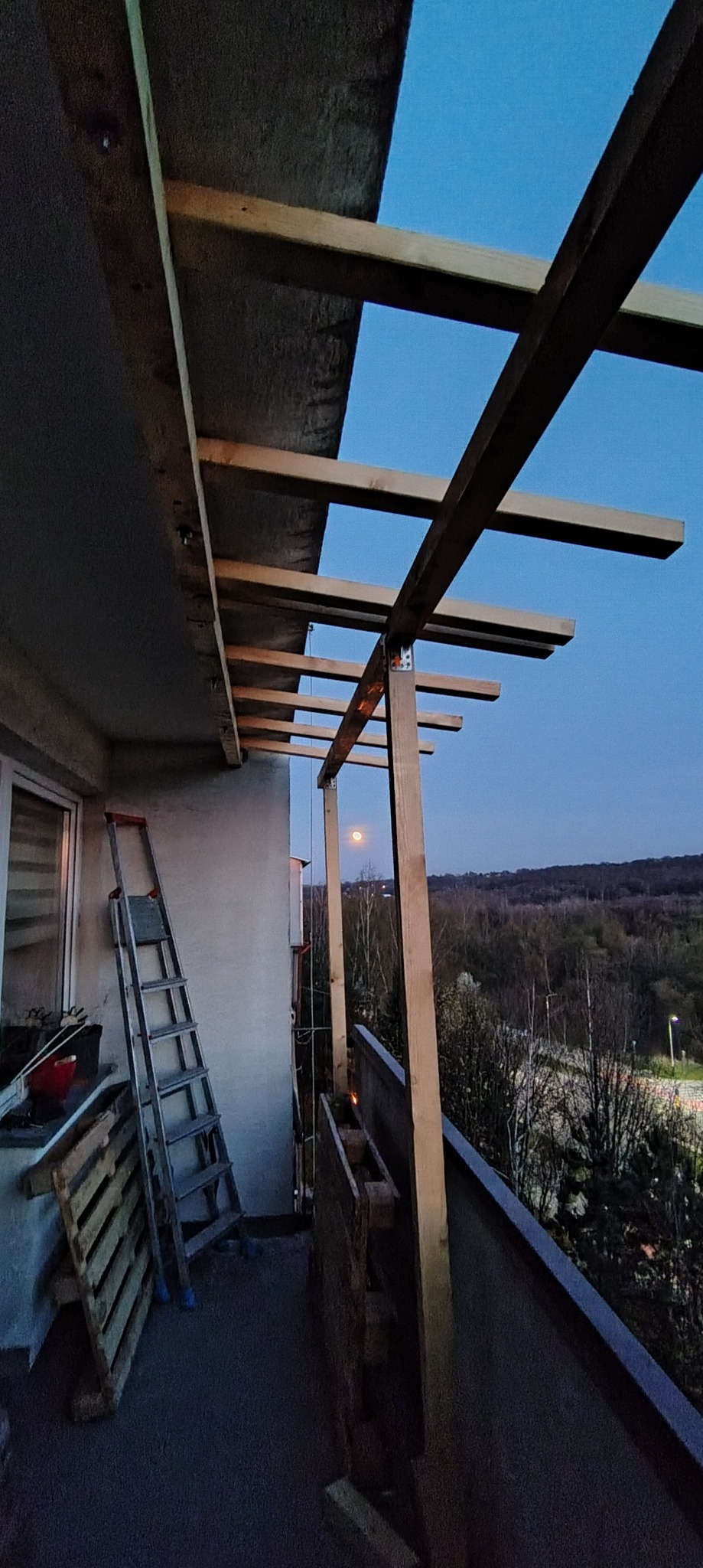 Konstrukcja drewniana zadaszenia na balkonie w trakcie montażu, widok na zewnątrz, drabina oparta o ścianę, wieczorny krajobraz z księżycem w tle.