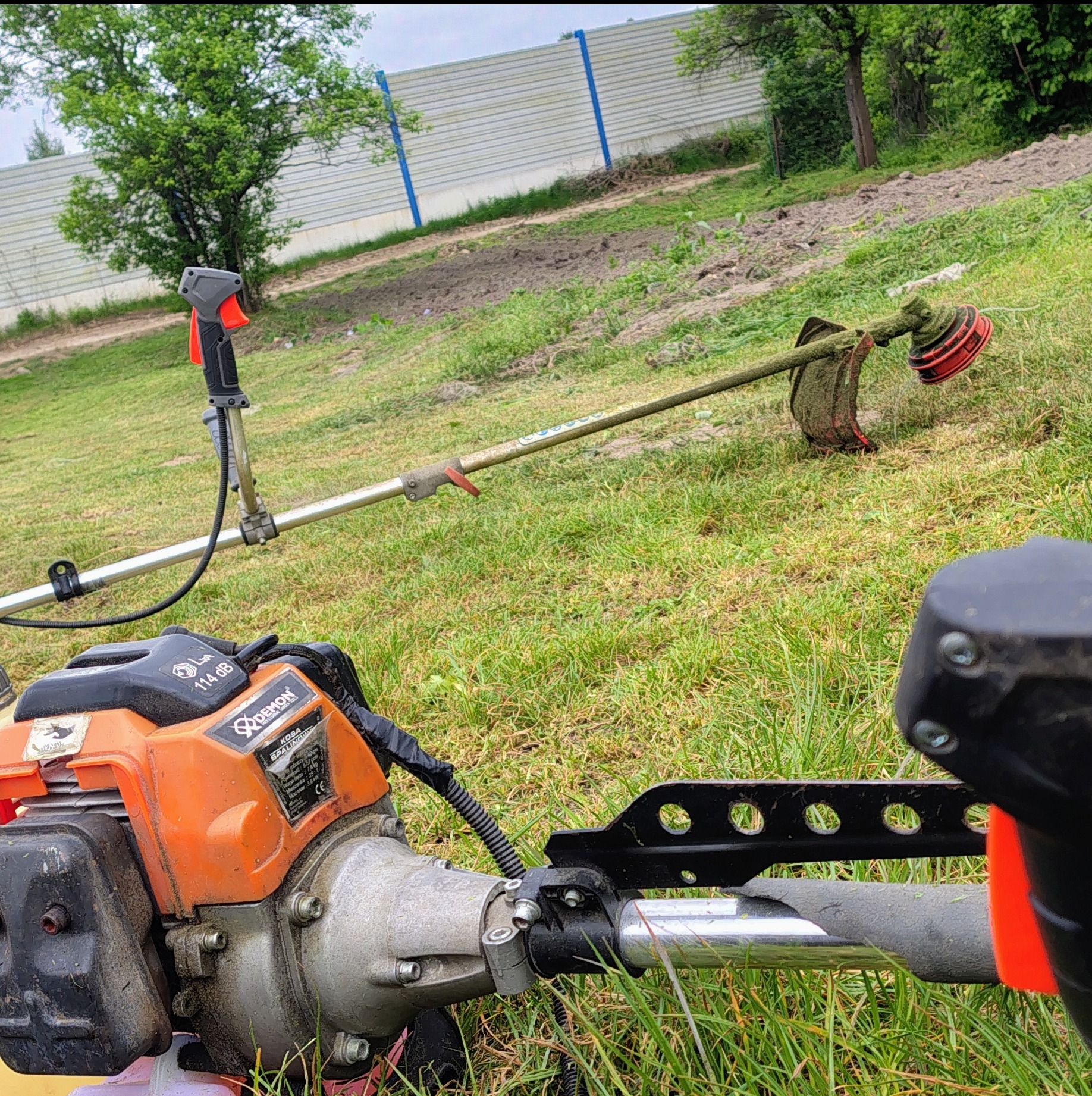Dwie kosiarki spalinowe na trawie, w tle ogrodzenie panelowe i drzewa. Jedna kosiarka leży na trawie, druga stoi oparta o nią. Widoczne ślady koszenia.