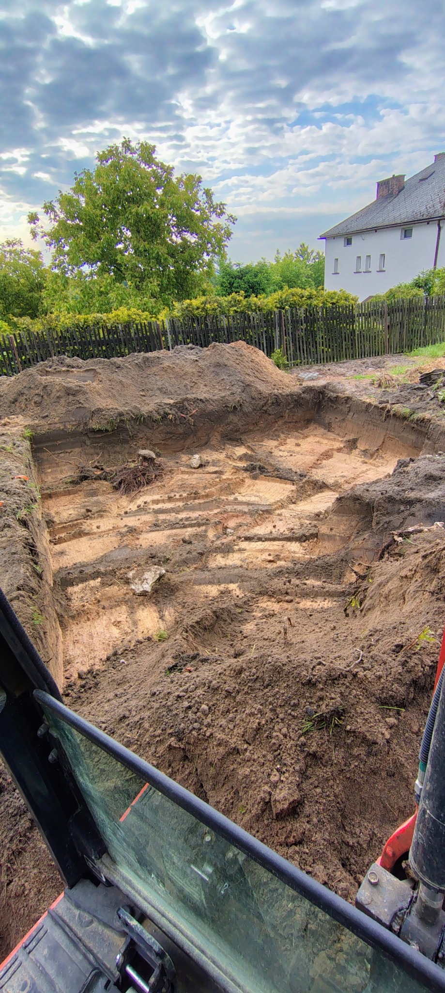 Wykonany wykop pod fundamenty domu jednorodzinnego, widok z kabiny koparki. W tle zielone drzewa i budynek mieszkalny.