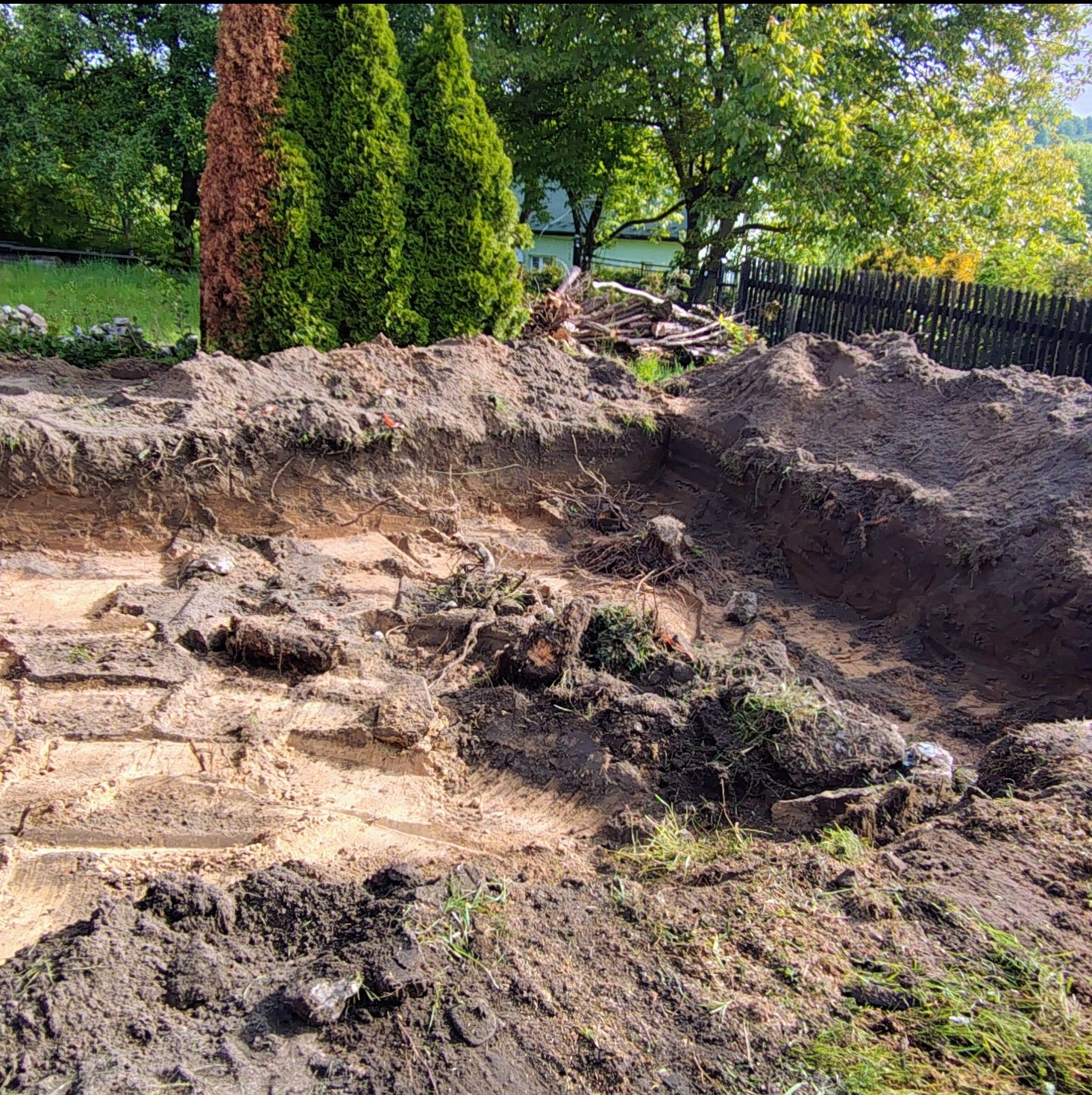 Wyrobisko w ziemi z widocznymi śladami opon, korzeniami i fragmentami darni. W tle drzewa i drewniany płot. Prace ziemne w trakcie realizacji.