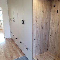 KAMIL CZYPIOR - Biała zabudowa meblowa z szafkami i szufladami, z okrągłymi czarnymi uchwytami, połączona z wnęką wykończoną drewnem z siedziskiem i oświetleniem LED, na pograniczu parkietu i szarej podłogi.