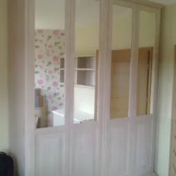 Jasna szafa wnękowa z lustrzanymi drzwiami, odbijająca wnętrze pokoju z tapetą w kwiaty i biurkiem