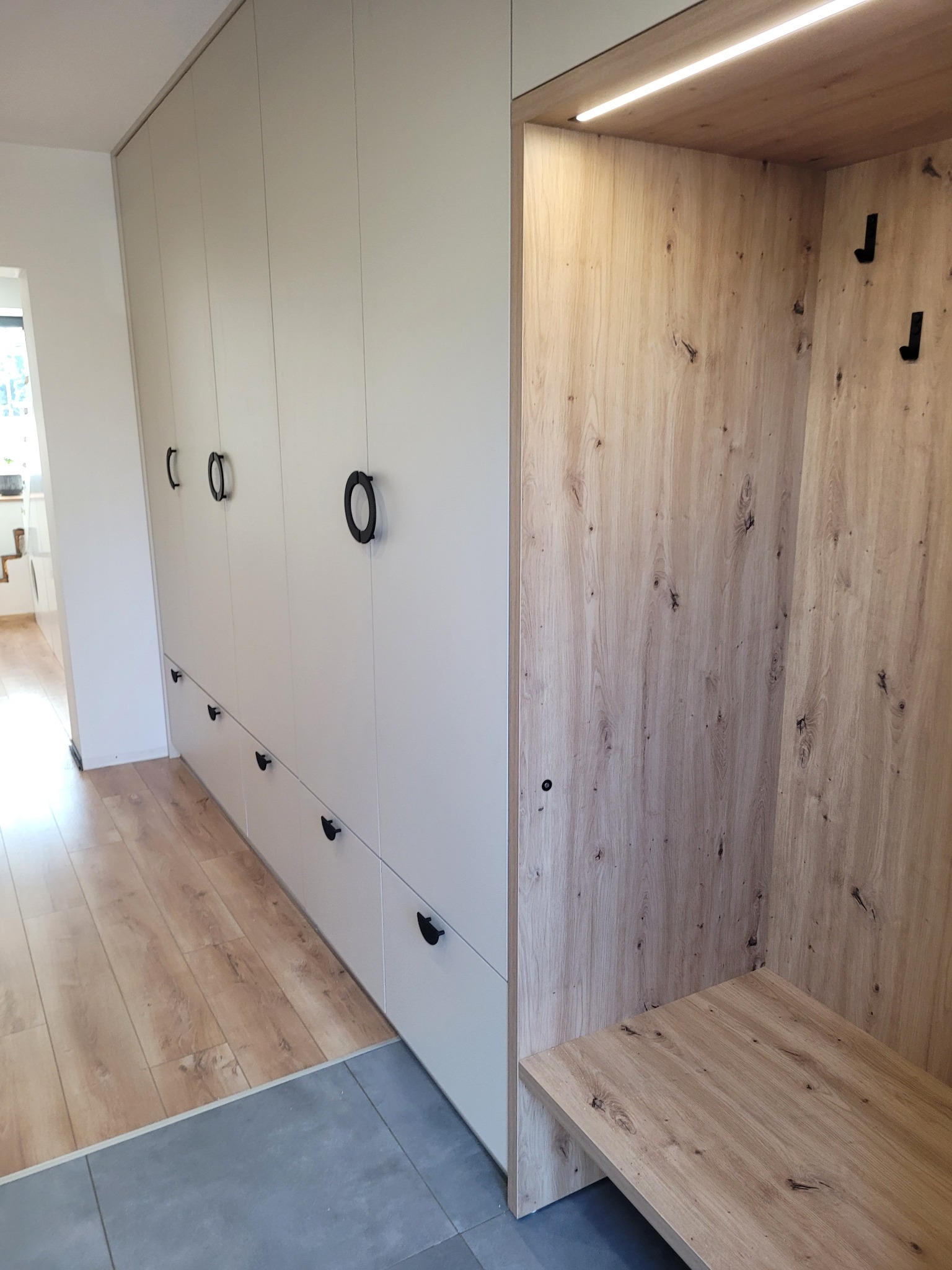 Biała zabudowa meblowa z szafkami i szufladami, z okrągłymi czarnymi uchwytami, połączona z wnęką wykończoną drewnem z siedziskiem i oświetleniem LED, na pograniczu parkietu i szarej podłogi.