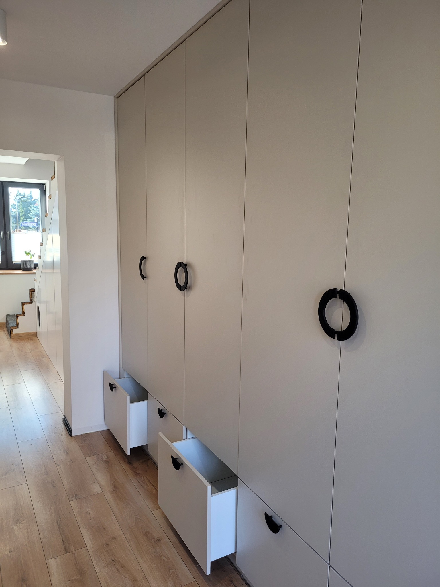 Jasnoszara zabudowa meblowa z szafkami do sufitu i wysuwanymi szufladami, czarne okrągłe uchwyty, podłoga drewnopodobna, widok na korytarz ze schodami w tle.