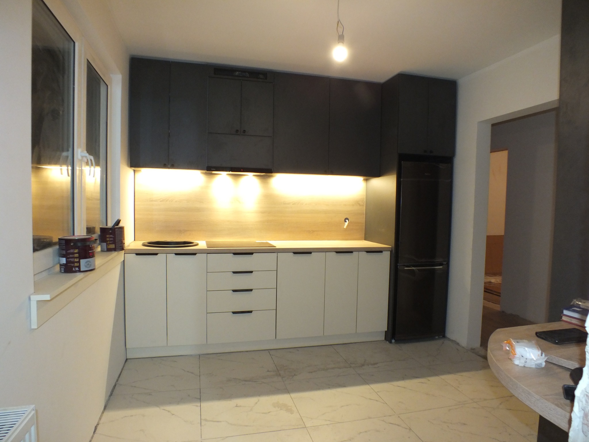 Nowoczesna kuchnia z białymi szafkami dolnymi i grafitowymi górnymi, podświetlana listwą LED, z czarną lodówką i marmurową podłogą, widoczne puszki z farbą na parapecie.