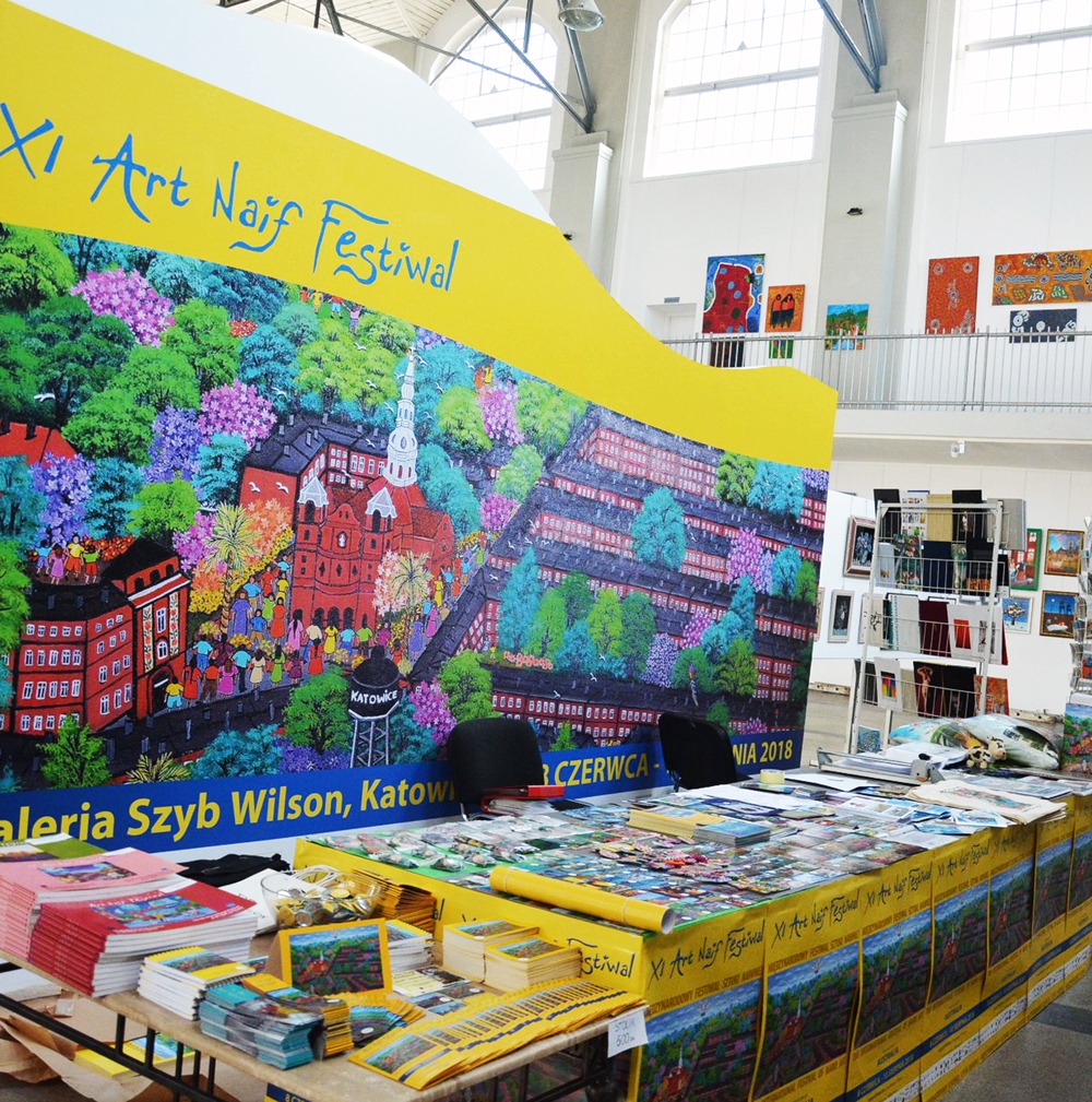 Stoisko wystawiennicze na XI Art Naif Festiwal w Galerii Szyb Wilson w Katowicach, z dużym banerem przedstawiającym kolorową, naiwną grafikę miejską i stosem materiałów promocyjnych na stole.