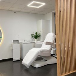 Instytut Urody Alicja Nikolin - Nowoczesny gabinet kosmetyczny z białym fotelem zabiegowym przykrytym jednorazowym pokrowcem, widoczny sprzęt kosmetyczny i drewniana ażurowa ścianka działowa.