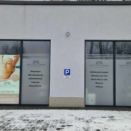 Instytut Urody Alicja Nikolin - Fasada budynku Instytutu Urody z dwoma dużymi oknami wystawowymi. Na szybach okien widoczne reklamy salonu, oferowane zabiegi kosmetyczne i numer telefonu. Nad wejściem znak parkingu dla klientów.