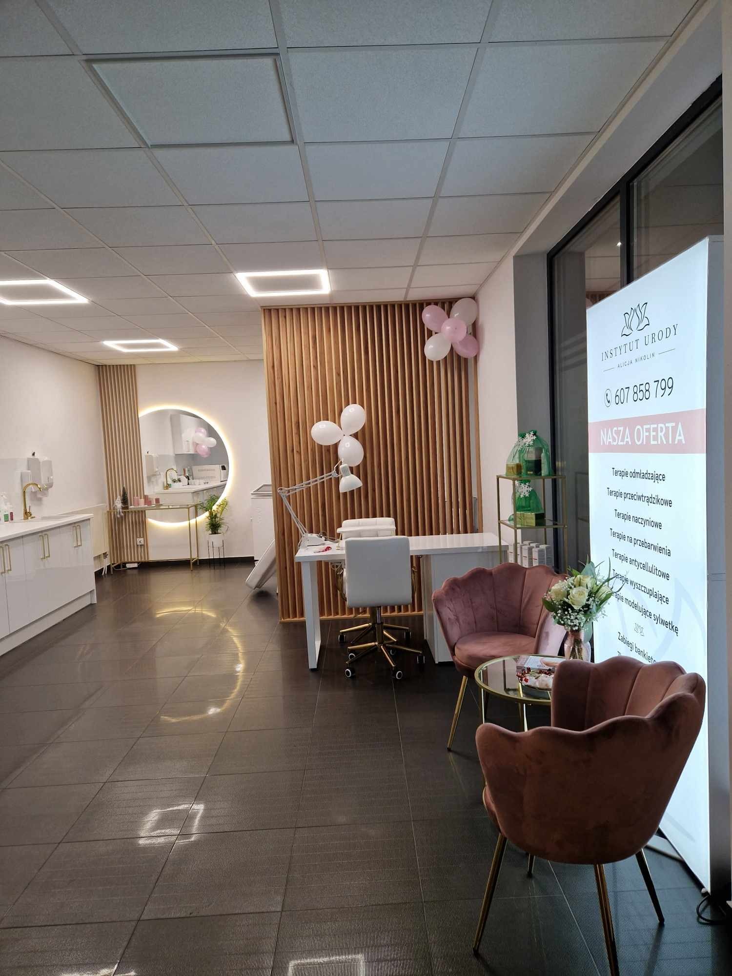 Jasne wnętrze salonu kosmetycznego z drewnianymi elementami dekoracyjnymi, stanowiskiem do manicure z lampą, okrągłym lustrem podświetlanym LED, komfortowymi fotelami w odcieniach różu i standem...