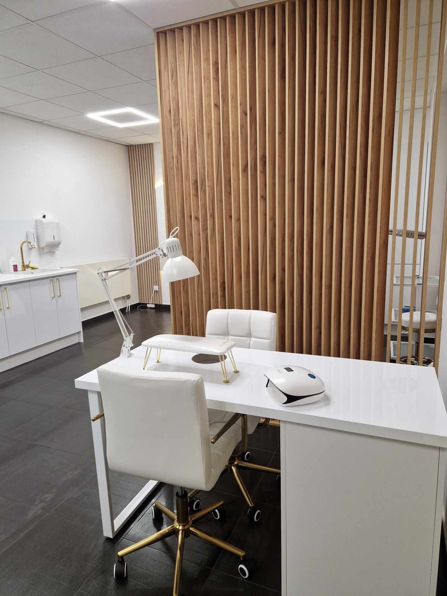 Nowoczesne stanowisko do manicure z białym biurkiem, lampą, pochłaniaczem pyłu i krzesłami na złotych nóżkach, oddzielone drewnianą ścianką działową w salonie kosmetycznym.