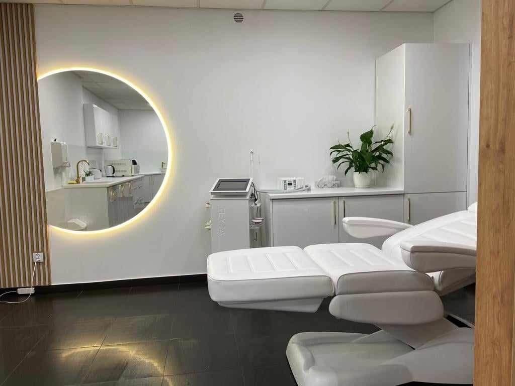 Jasne wnętrze gabinetu kosmetycznego z białym, regulowanym fotelem do zabiegów, oświetlonym lustrem w kształcie półksiężyca i sprzętem kosmetycznym. W tle widoczny fragment pomieszczenia z umywalką.