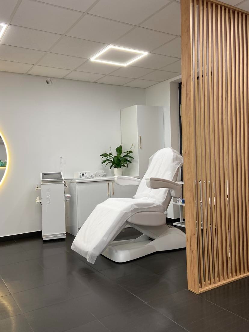 Nowoczesny gabinet kosmetyczny z białym fotelem zabiegowym przykrytym jednorazowym pokrowcem, widoczny sprzęt kosmetyczny i drewniana ażurowa ścianka działowa.