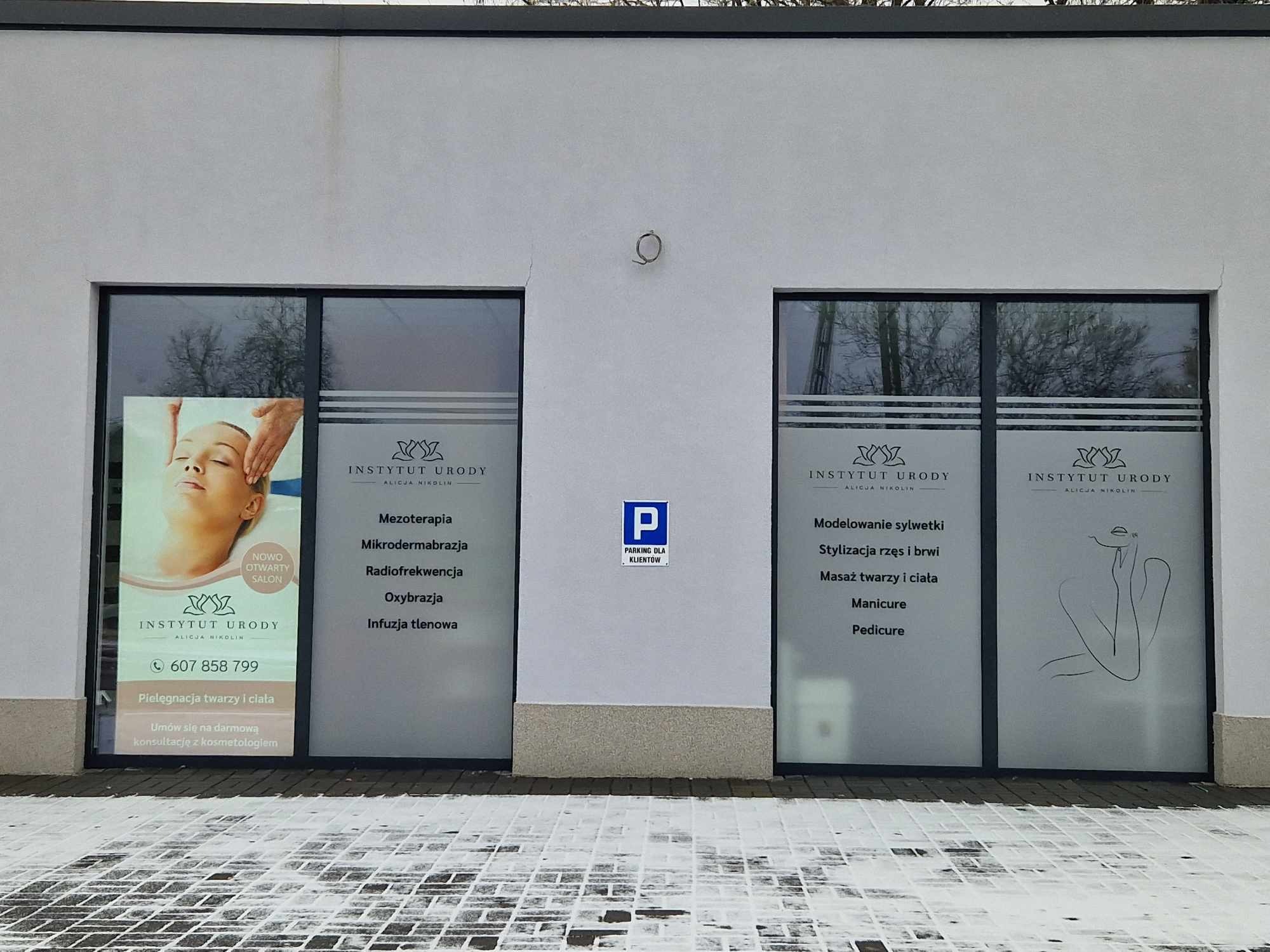Fasada budynku Instytutu Urody z dwoma dużymi oknami wystawowymi. Na szybach okien widoczne reklamy salonu, oferowane zabiegi kosmetyczne i numer telefonu. Nad wejściem znak parkingu dla klientów.