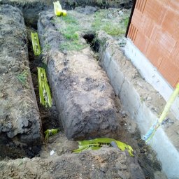 Inbuko Sp. z o.o. - Wykopane rowy pod fundamenty budynku z widocznymi żółtymi rurami drenarskimi w folii ochronnej, częściowo zasypane ziemią, z fragmentem ściany budynku w tle.