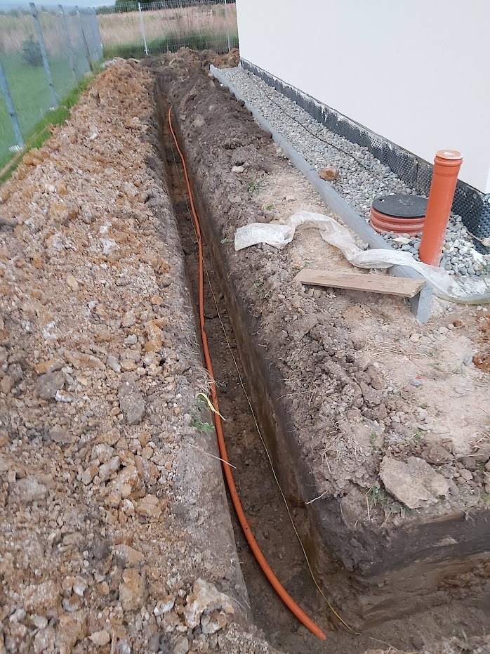 Wykonany wykop pod rurę kanalizacyjną wzdłuż fundamentu budynku, widoczna pomarańczowa rura ułożona w wykopie, obok rury widoczna linka wyznaczająca trasę.
