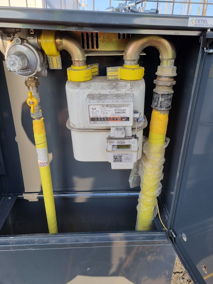 Zamknięta szafka gazowa z licznikiem Intergaz, żółte rury gazowe owinięte taśmą, widoczne złącza i zawór.