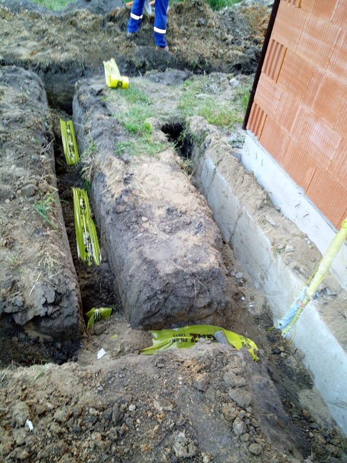 Wykopane rowy pod fundamenty budynku z widocznymi żółtymi rurami drenarskimi w folii ochronnej, częściowo zasypane ziemią, z fragmentem ściany budynku w tle.