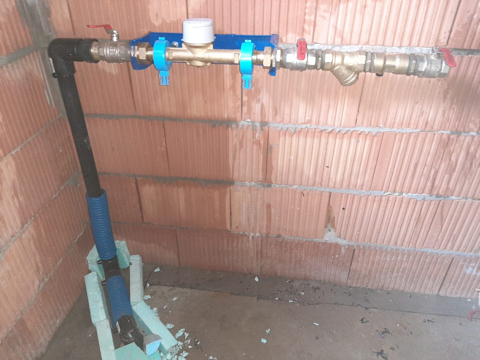 Nowe przyłącze wodociągowe z licznikiem zamontowane na ścianie z cegły, rura w niebieskiej osłonie wsparta na styropianowych blokach.