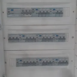 Rozdzielnica elektryczna z bezpiecznikami i wyłącznikami, widoczne diody sygnalizacyjne, obudowa biała, skrzynka otwarta.