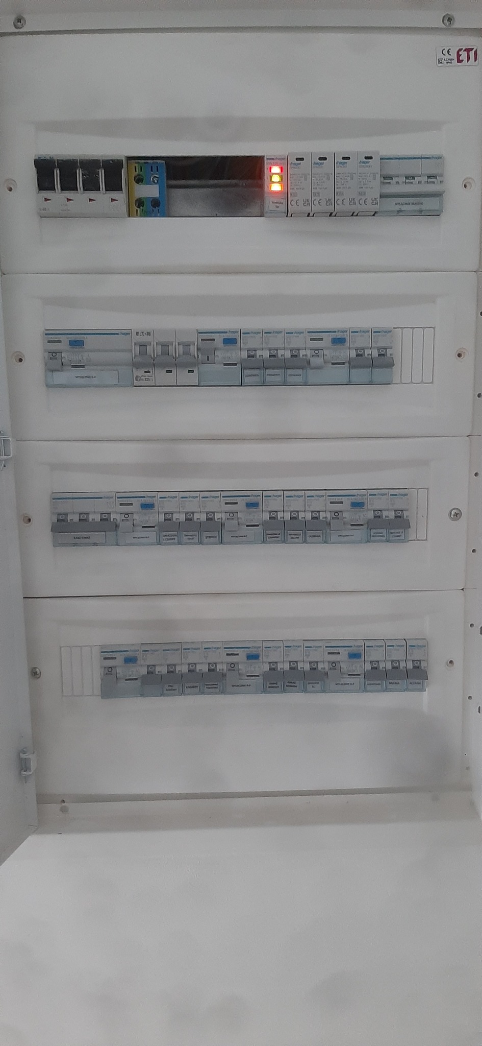 Rozdzielnica elektryczna z bezpiecznikami i wyłącznikami, widoczne diody sygnalizacyjne, obudowa biała, skrzynka otwarta.