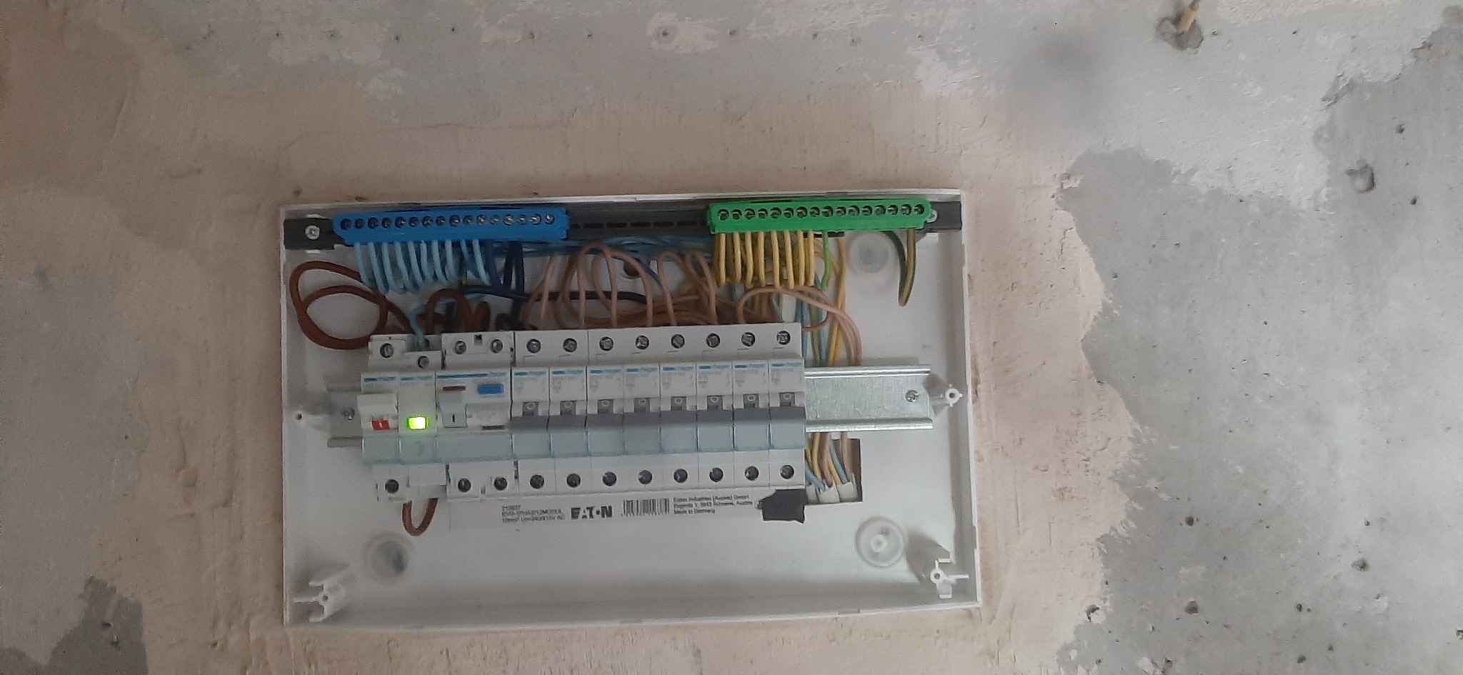 Rozdzielnica elektryczna Eaton zamontowana na surowej ścianie, widoczne przewody, listwy zaciskowe i zabezpieczenia, jeden z wyłączników z sygnalizacją LED.