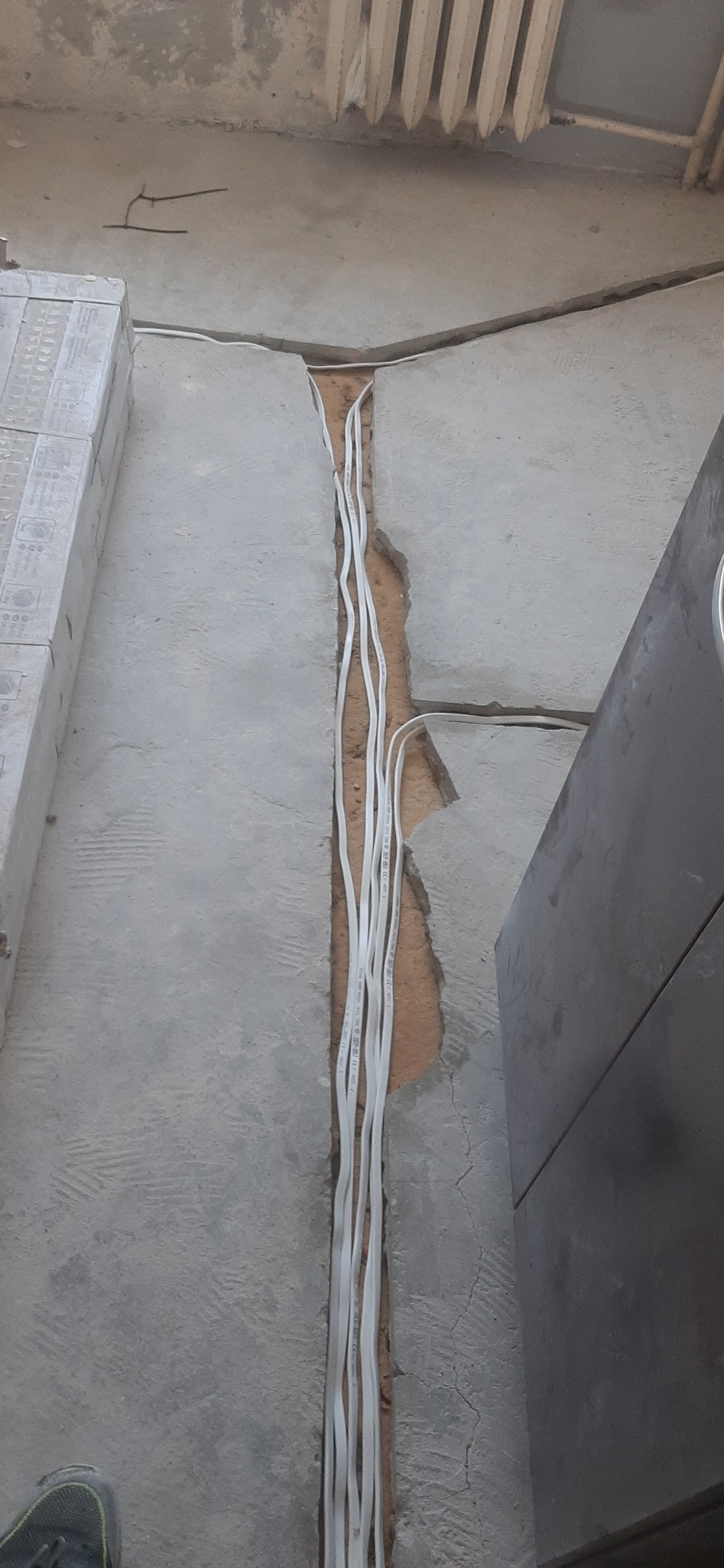 Bruzda w szarej, betonowej posadzce z ułożonymi w niej przewodami elektrycznymi w białej izolacji, widok z góry, fragment grzejnika i buta w kadrze.