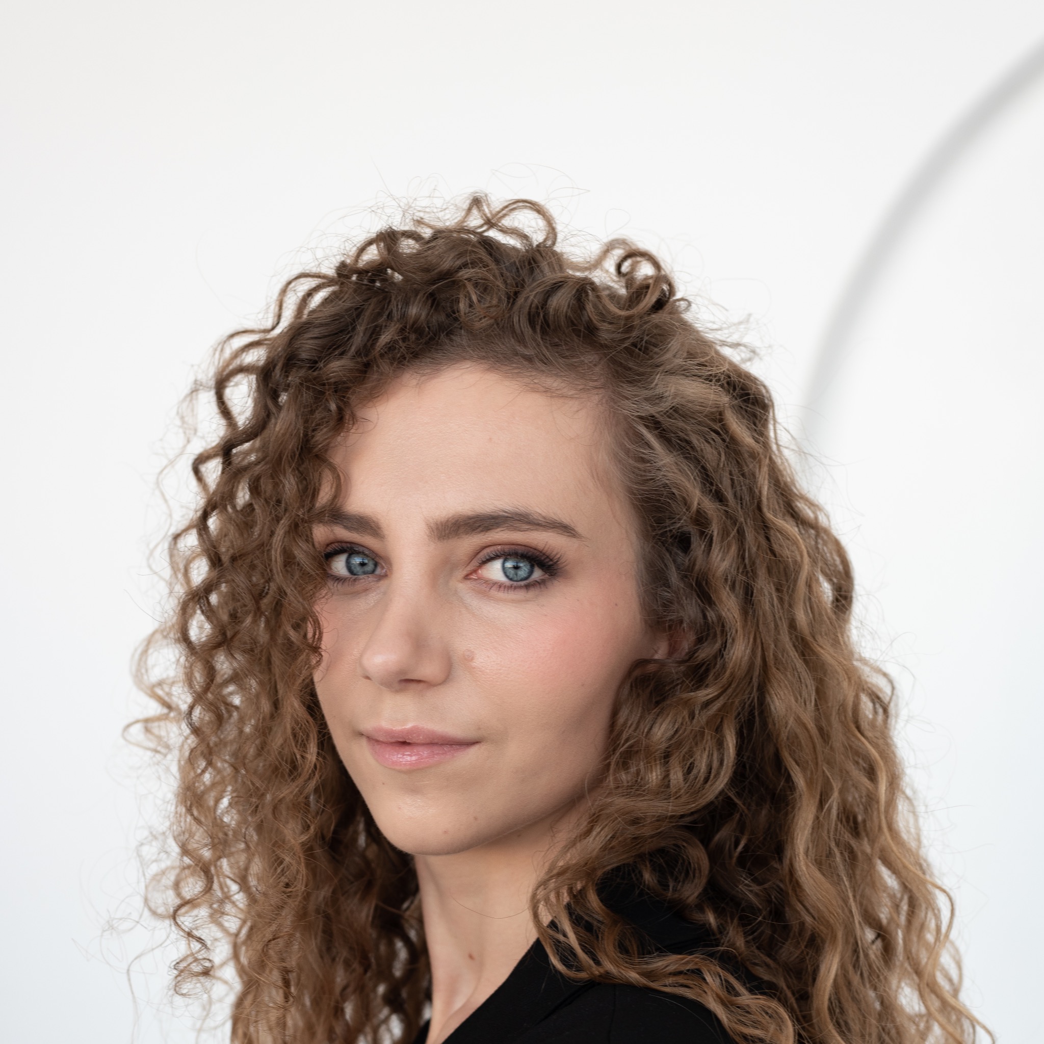 Portret kobiety o niebieskich oczach i kręconych brązowych włosach, ubrana w czarną marynarkę na jasnym tle.