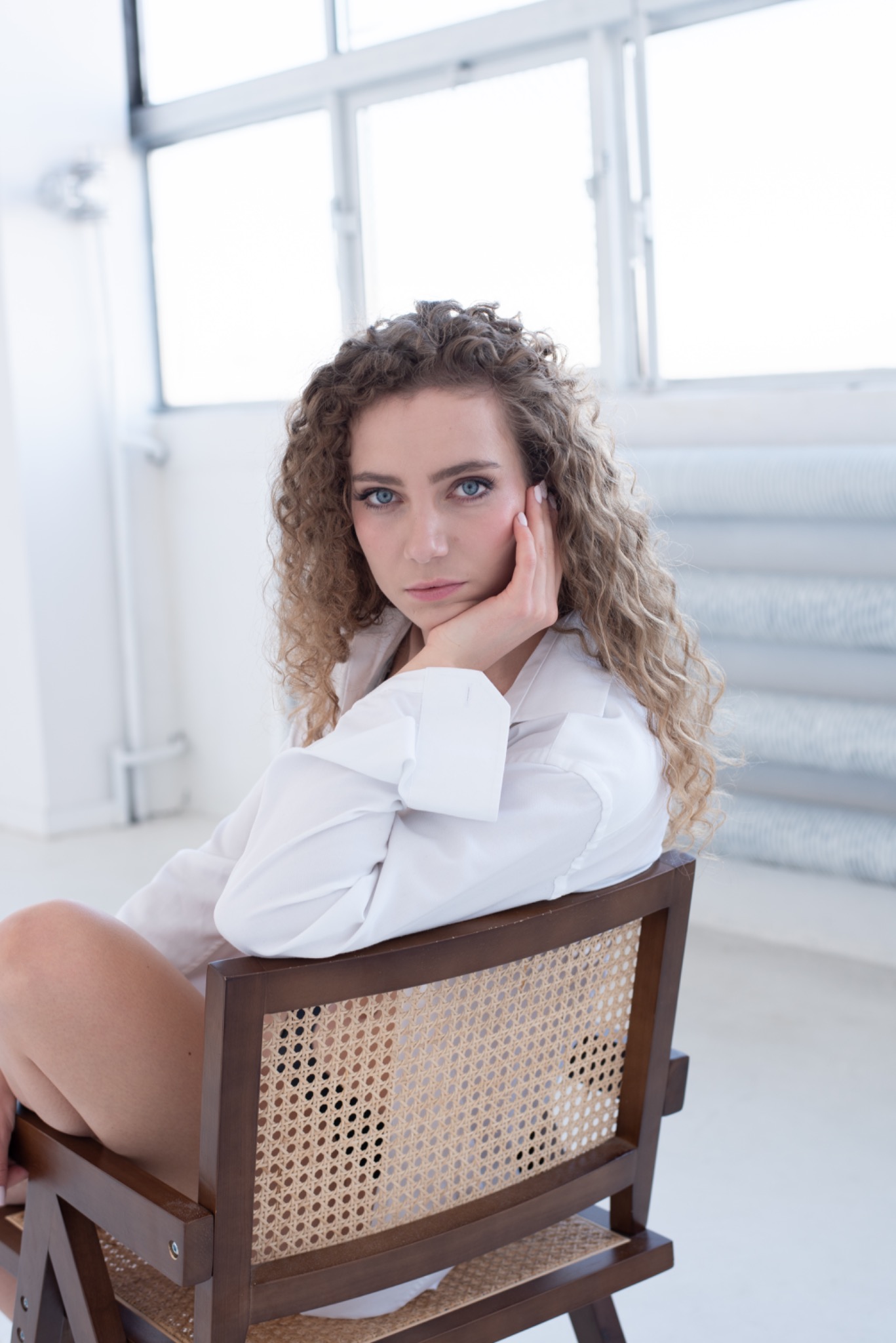 Portret kobiety z kręconymi włosami w białej koszuli, opierającej brodę na dłoni, siedzącej na krześle z plecionym oparciem, na tle jasnego wnętrza z oknem.