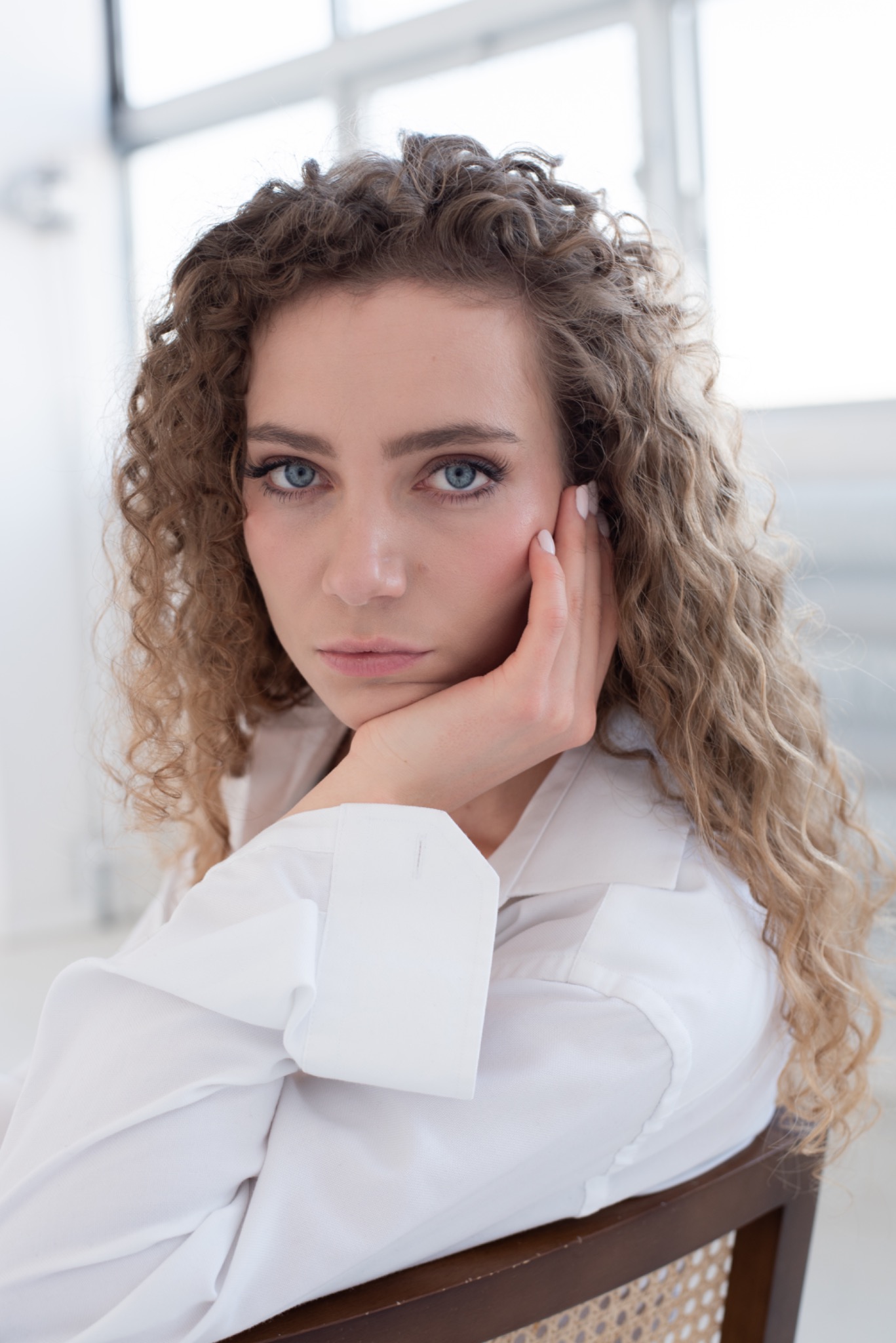 Portret kobiety o kręconych włosach i niebieskich oczach, w białej koszuli, opierającej policzek na dłoni, w jasnym, minimalistycznym wnętrzu.