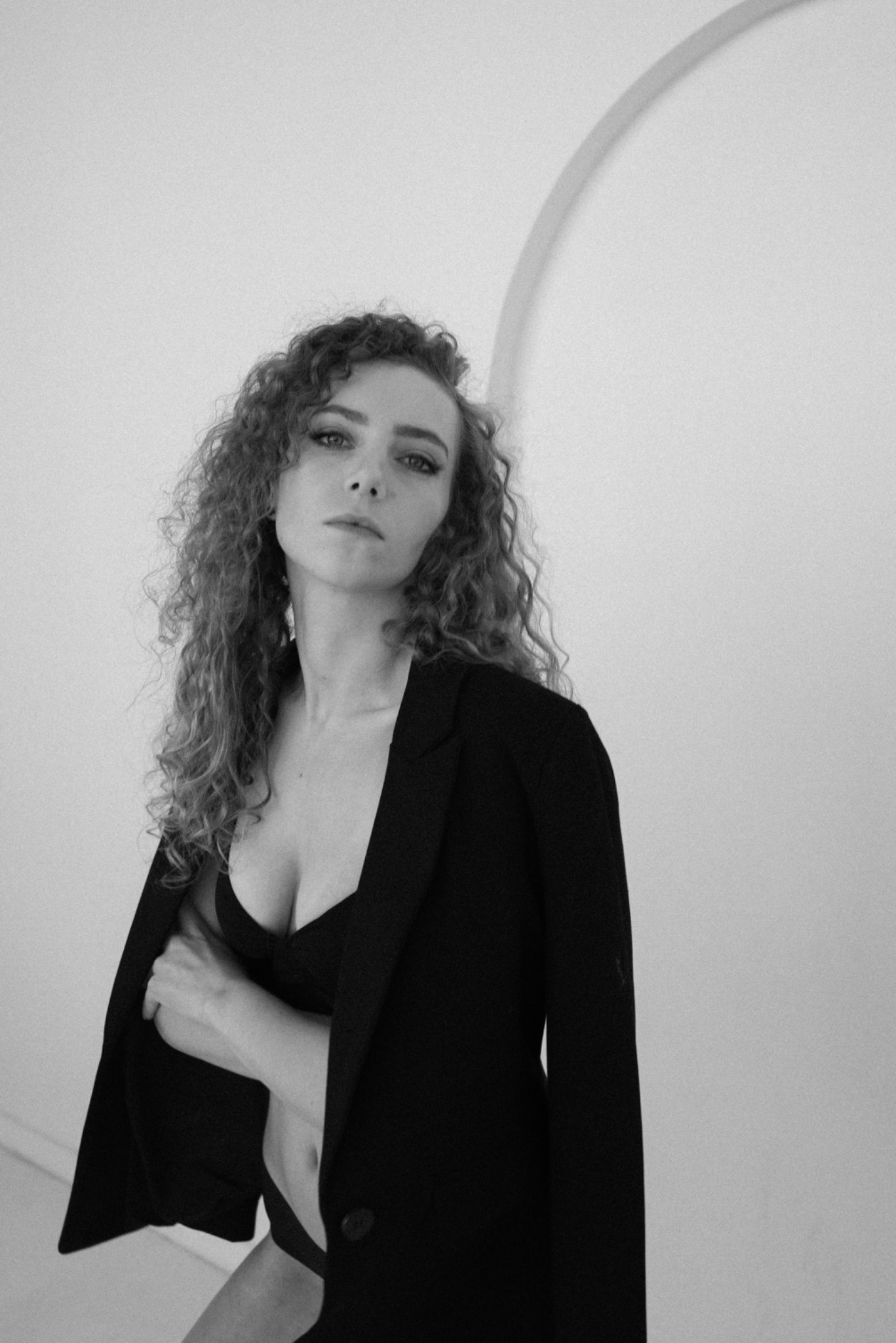 Portret kobiety z kręconymi włosami w czarnej marynarce i bieliźnie na tle minimalistycznego wnętrza z łukiem