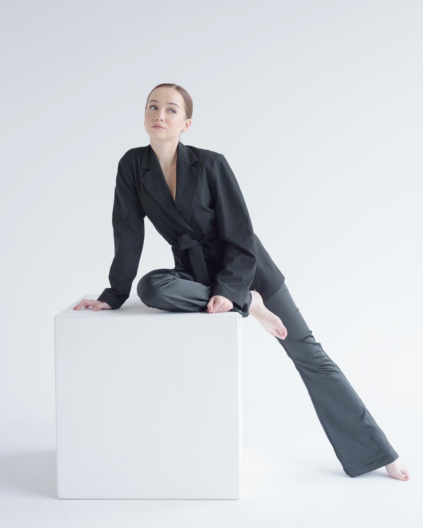 Kobieta w czarnym garniturze z paskiem, siedząca na białym sześcianie, z nogami wyciągniętymi w bok, patrzy w górę na jasnym tle.