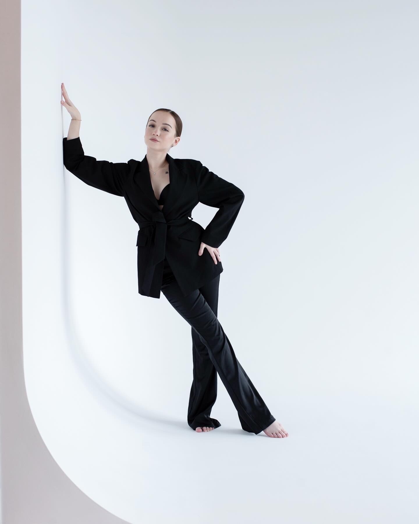 Kobieta w czarnym garniturze, pozująca boso na białym tle, opierająca się o ścianę, z delikatnym makijażem i włosami upiętymi w kok