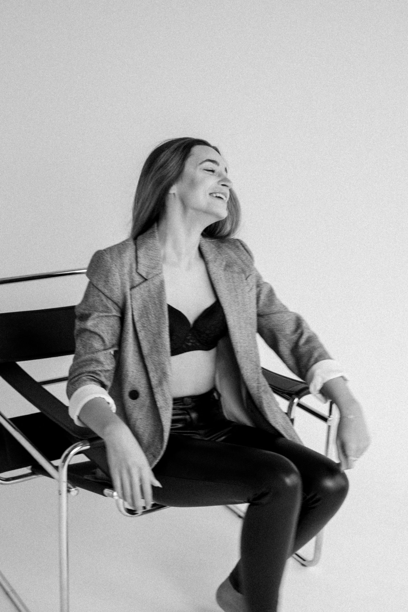 Czarno-białe zdjęcie uśmiechniętej kobiety siedzącej na krześle w marynarce i bieliźnie, z głową odchyloną do tyłu, na jasnym tle.