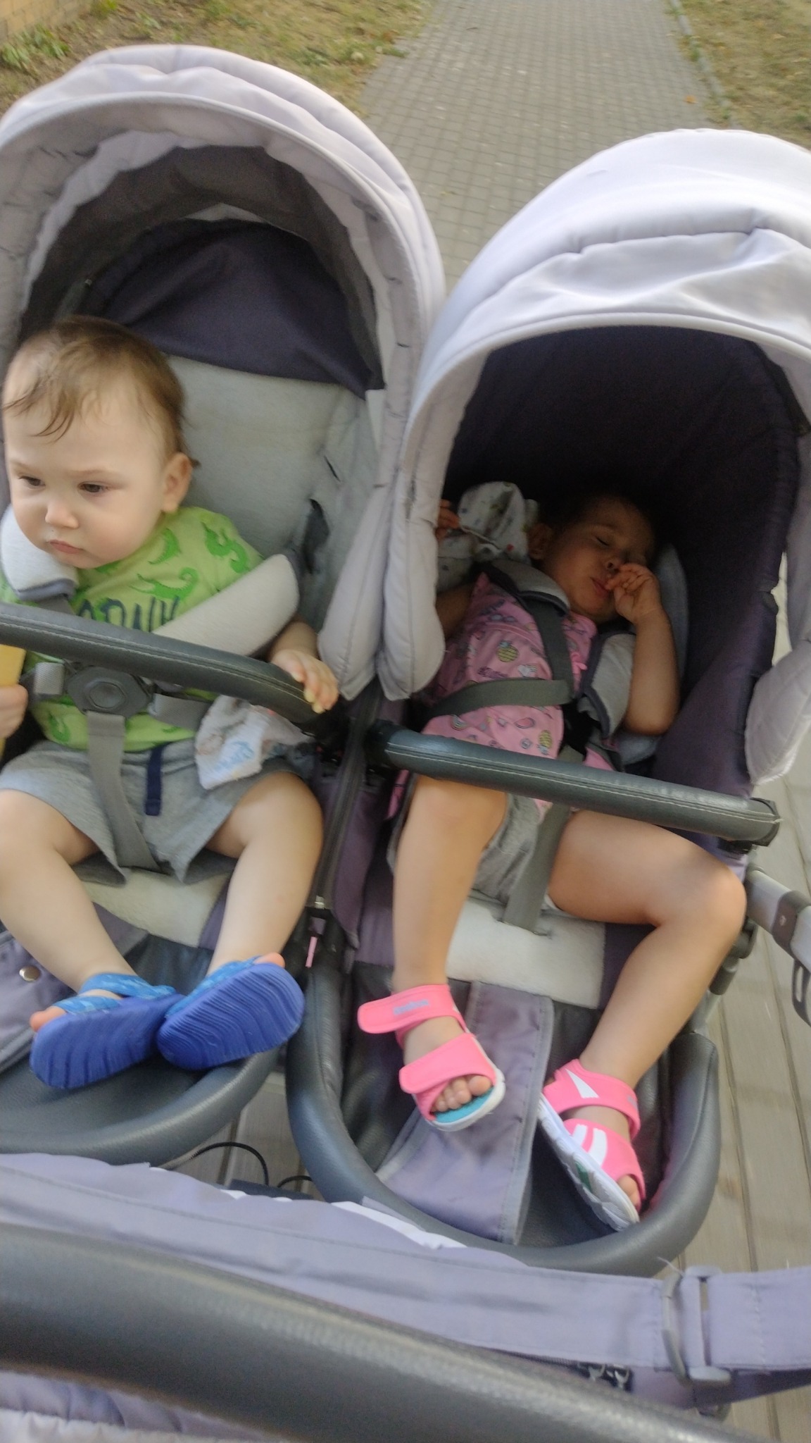 Dwoje dzieci w wózku bliźniaczym: jedno siedzi i trzyma przekąskę, drugie śpi z dłonią przy ustach.