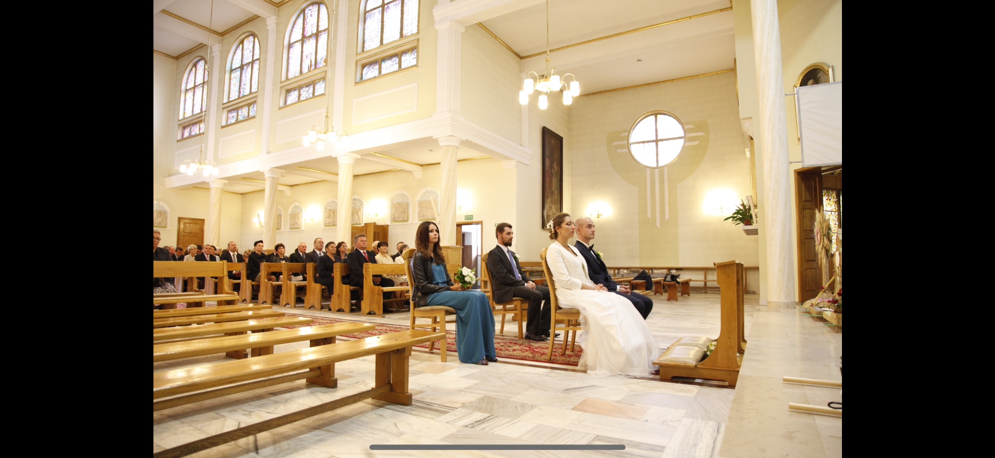 Para młoda siedzi w kościele podczas ceremonii ślubnej, w tle goście i drewniane ławki, widać dekoracje kwiatowe i jasne wnętrze kościoła z wysokimi oknami.
