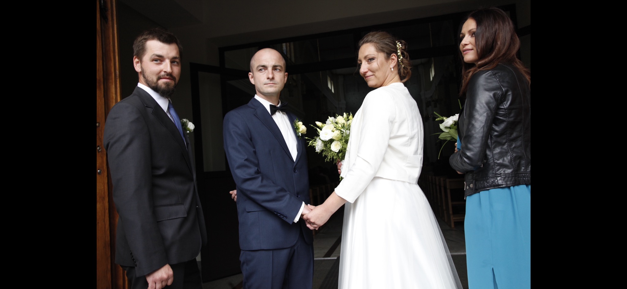 Para młoda trzymająca się za ręce przed kościołem w Warszawie, obok świadkowie, panna młoda w białej sukni i bolerku, pan młody w granatowym garniturze z muchą.