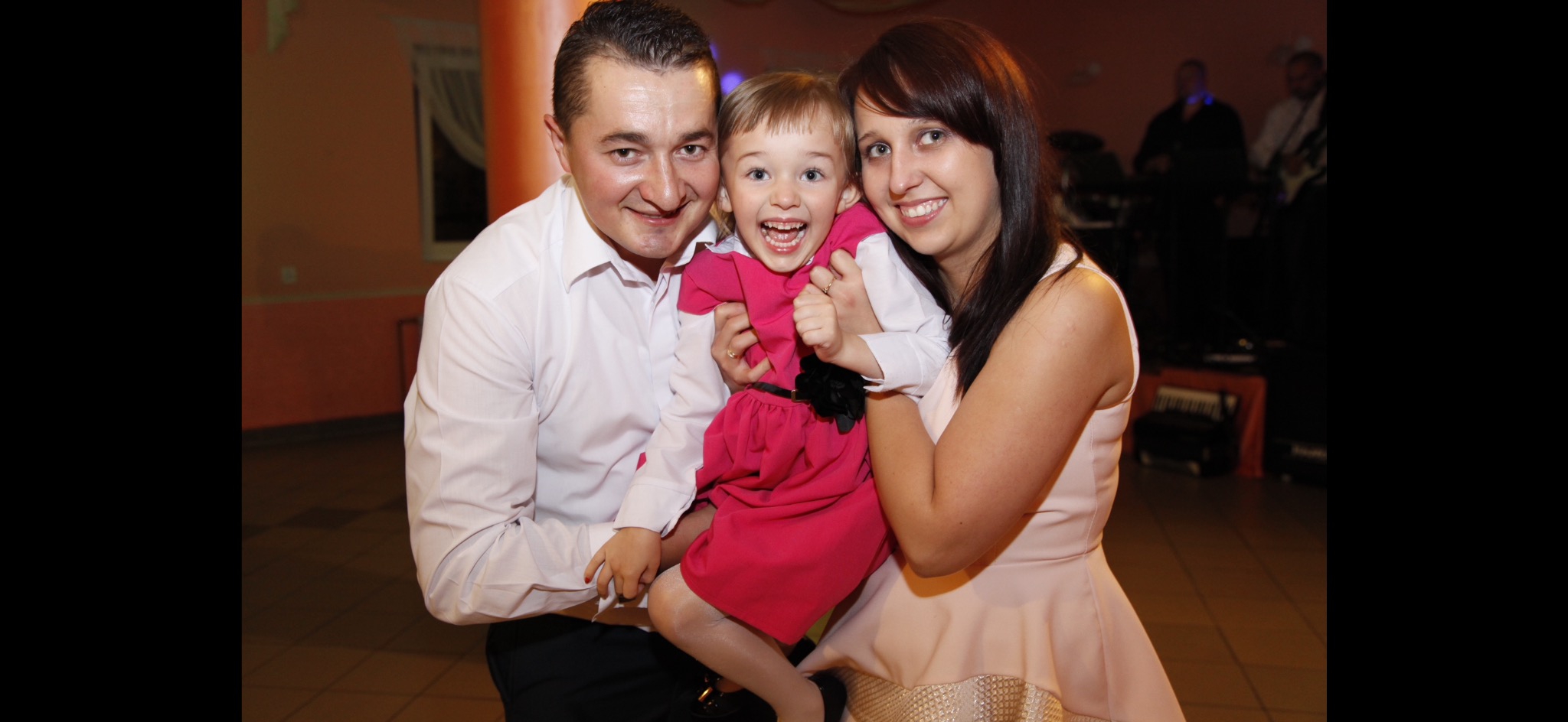 Uśmiechnięta rodzina: tata w białej koszuli trzyma córeczkę w różowej sukience, obok mama w jasnej sukience, w tle zespół muzyczny.