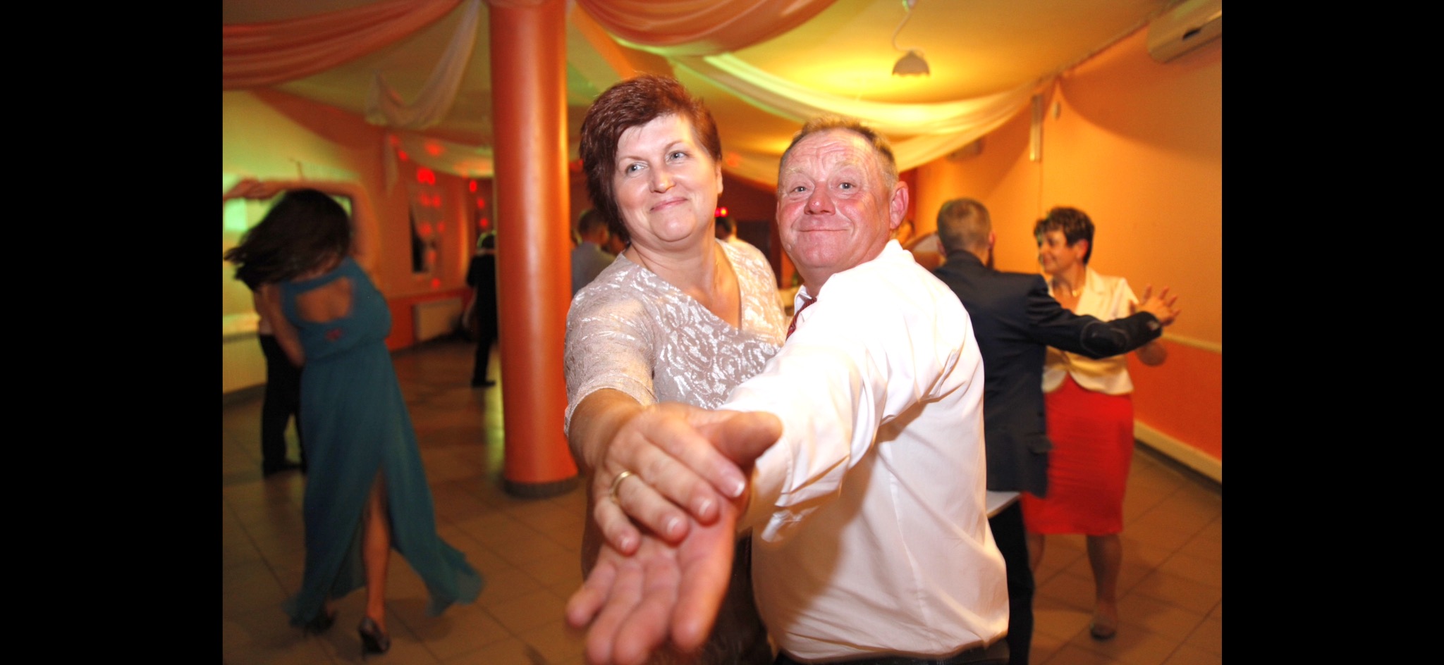 Uśmiechnięta para w trakcie pierwszego tańca weselnego, w tle tańczący goście weselni, sala udekorowana pomarańczowymi i białymi materiałami.