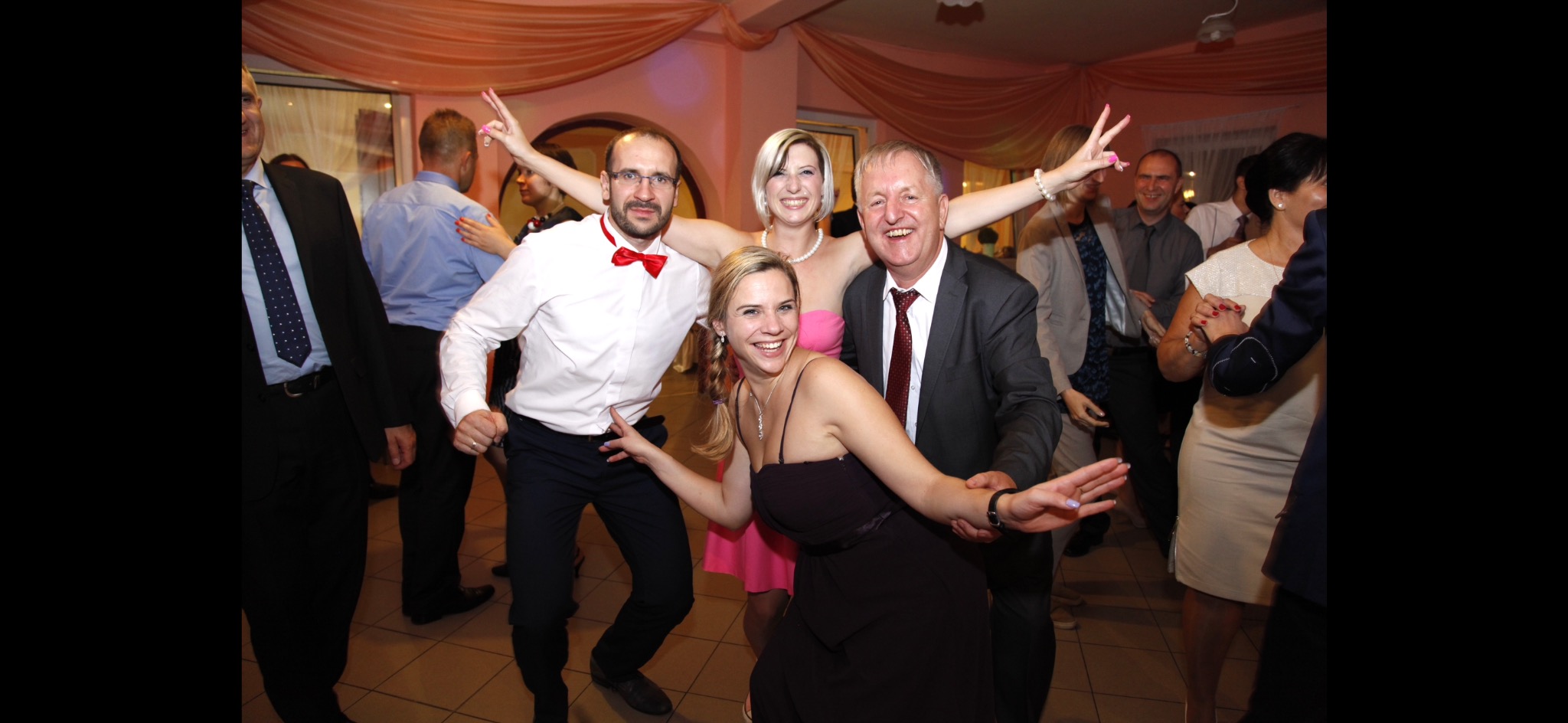 Rozbawieni goście weselni tańczą na sali, mężczyzna w okularach i z czerwoną muchą pokazuje znak zwycięstwa, tło stanowią dekoracje z materiału w kolorze brzoskwiniowym.