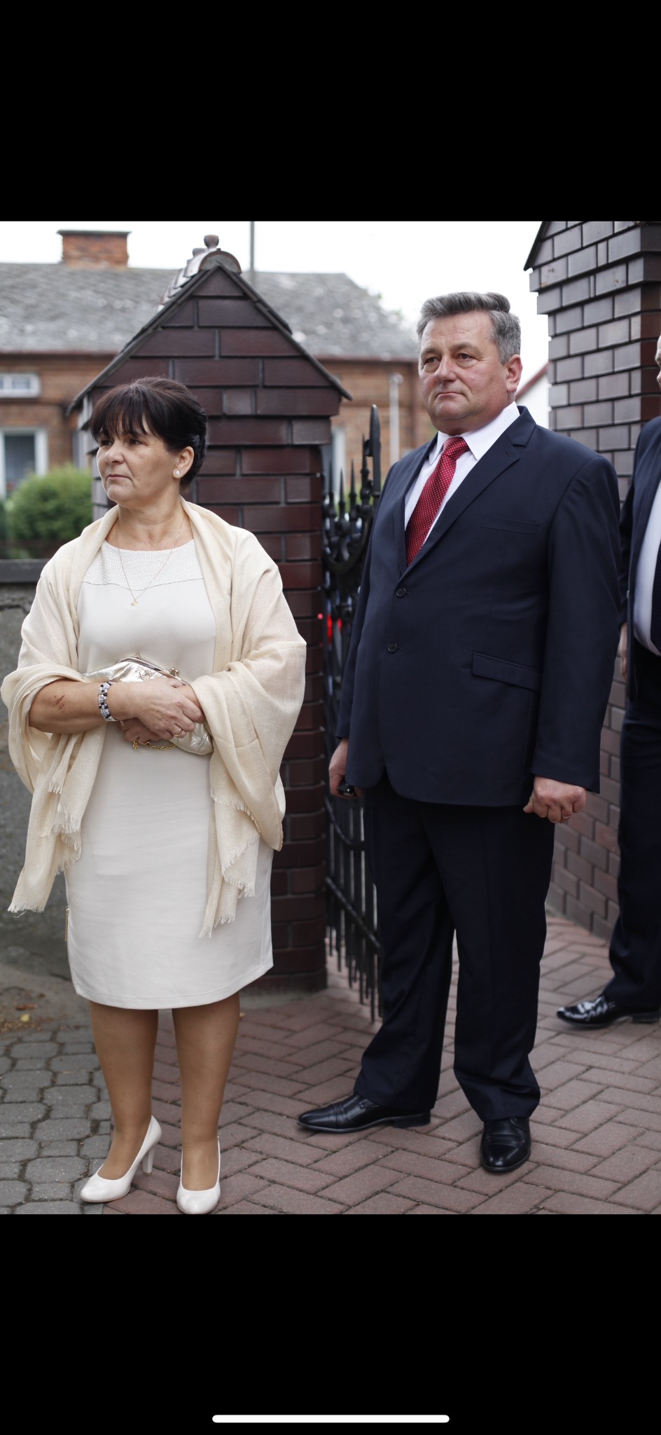 Elegancko ubrana para, kobieta w kremowej sukience i szalu, mężczyzna w granatowym garniturze z czerwonym krawatem, pozują na tle ceglanego muru i metalowej bramy.
