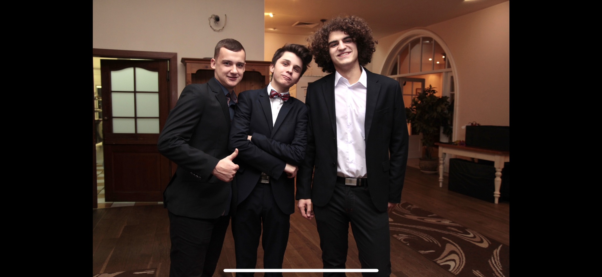 Trzech młodych mężczyzn w garniturach pozuje do zdjęcia w eleganckim wnętrzu z drewnianą podłogą i dekoracyjnym dywanem. Jeden z mężczyzn pokazuje kciuk w górę.