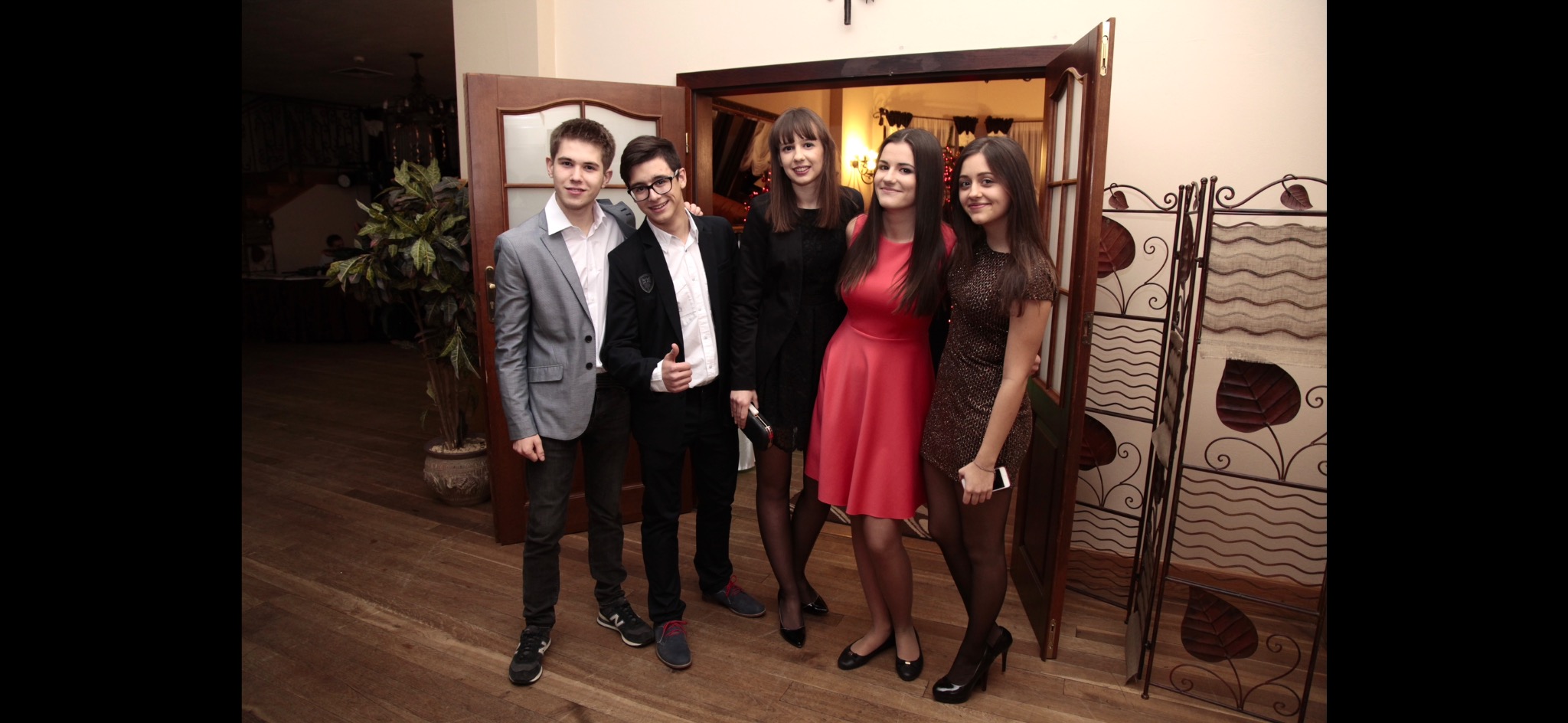 Grupa pięciu młodych osób, trzech dziewcząt w sukienkach i dwóch chłopców w marynarkach, pozuje do zdjęcia w pomieszczeniu z drewnianą podłogą i dekoracyjnym parawanem.