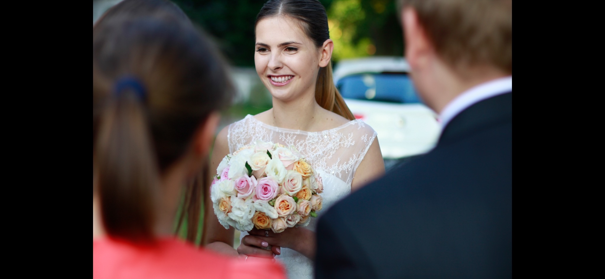 Uśmiechnięta panna młoda w koronkowej sukni trzyma bukiet róż w odcieniach bieli, różu i brzoskwini podczas ceremonii ślubnej, widoczna z perspektywy gości.